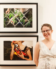 Dr. Ivana George smiling with brown hair pulled up wearing black rim glasses and a sleeveless v-neck off white dress standing beside 2 framed photographs on display