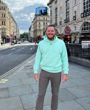 Dr. Andrew Miller standing on a city sidewalk smiling with reddish brown short hair, mustache and beard wearing a light blue green hoodie sweatshirt and gray pants